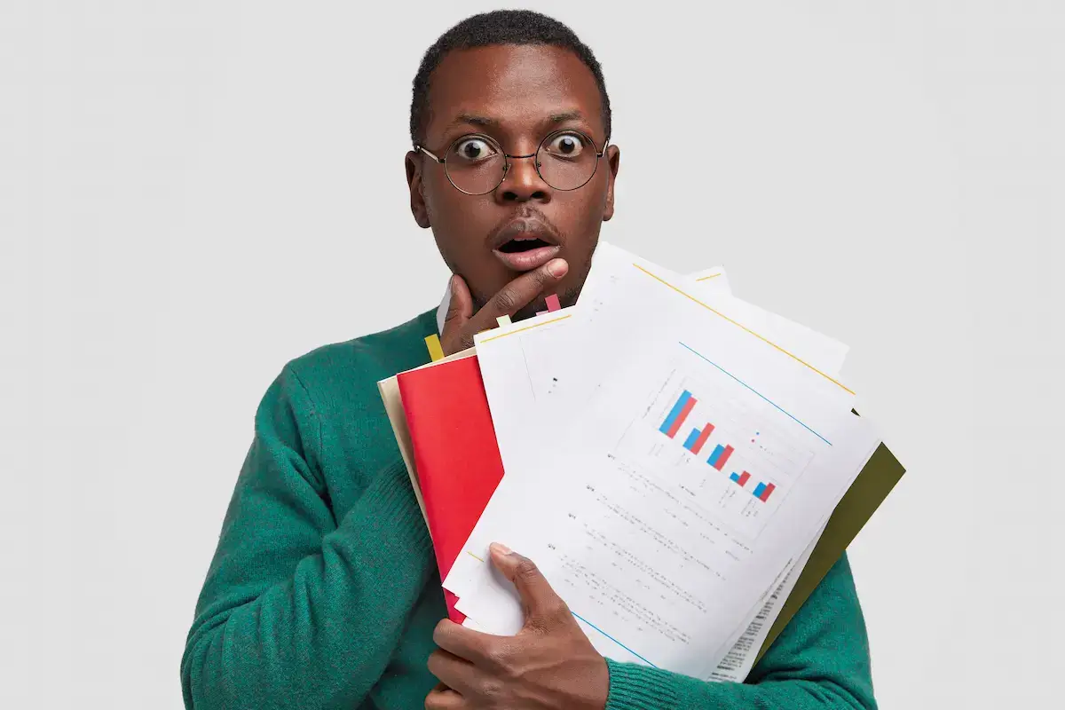 Surprised man wearing glasses and green sweater holding papers with charts and graphs against a plain background.